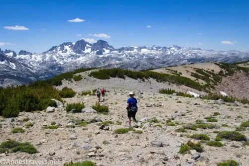 Three hikers on a rocky ridgetop with bushes and snowy peaks in the distance