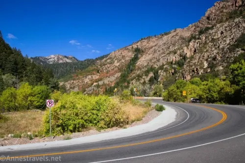 A curve in the paved Big Cottonwood Canyon Road with yellow lines down the middle, bordered by white concrete, no parking signs, and mountain cliffs 