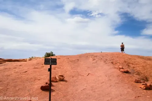 A signpost on slickrock for the first Upheaval Dome Overlook and a hiker against clouds in a blue sky 