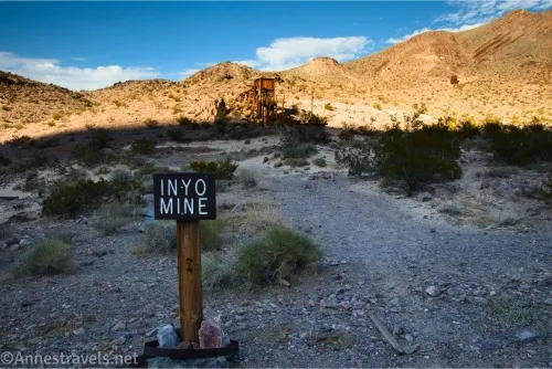 A wooden sign that read Inyo Mine in front of a desert and gravel path, a wooden structure, and distant hills 
