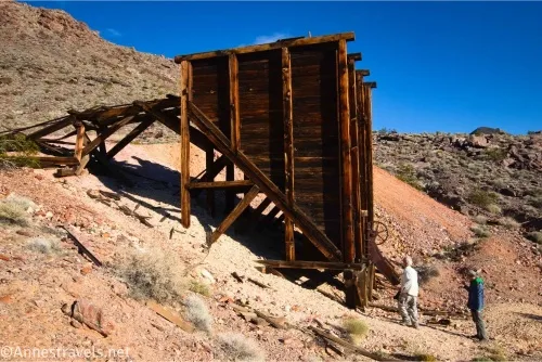 A large, wooden ore bin and two hikers on a desert hillside