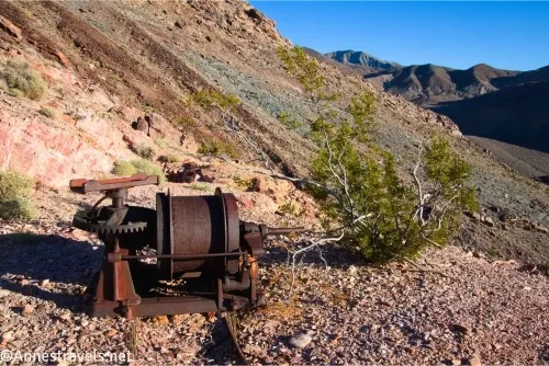 A rusty piece of machinery beside a greasebush on a desert hillside with mountains in the distance 