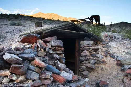 A cabin dug into the hillside with the front built of rocks, while in the distance is a wooden structure and mountains 