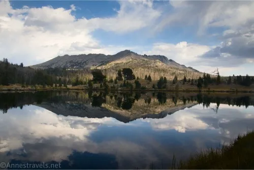 Reflections in a lake of clouds in a blue sky, a rugged mountain, and trees 