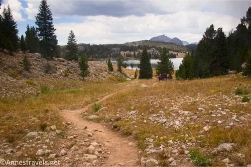 A trail in a grassy and rocky meadow with trees between the meadow and a lake and distant mountains below cloudy skies 
