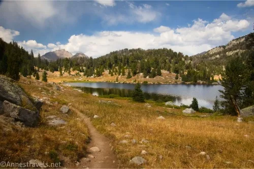A trail in a grassy meadow beside a lake with hills and mountains across the lake and clouds in the sky