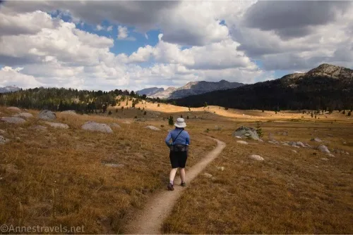 A hiker on a path in grassy meadows beneath cloudy skies with patches of blue and distant hills and mountains 