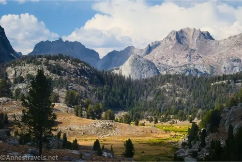 A rugged mountain ridgeline against clouds with a closer forested hill and meadows 