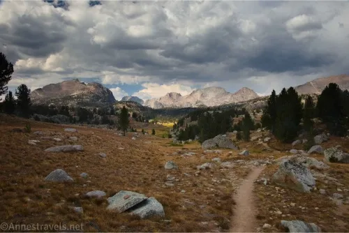 A trail in a shadowed, rocky meadow with distant rugged mountains and clouds overhead