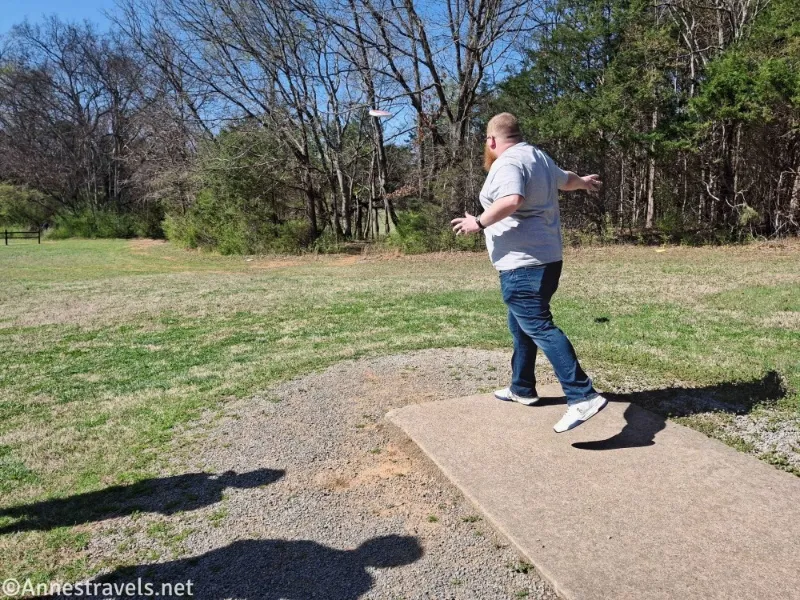A man stands on a concrete pad throwing a frisbee into a grassy area and trees