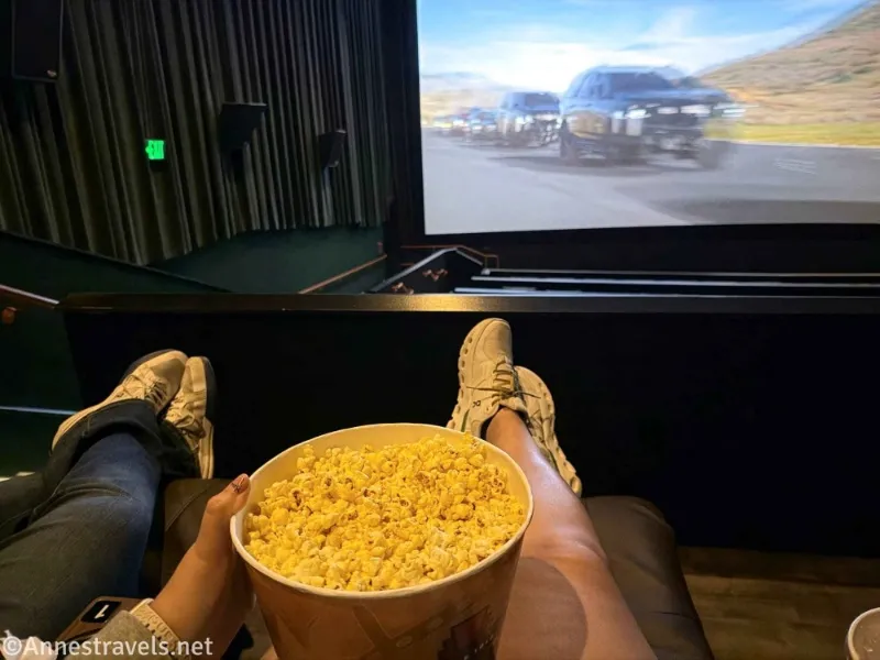A large bucket of popcorn in front of a movie screen with cars on the screen 