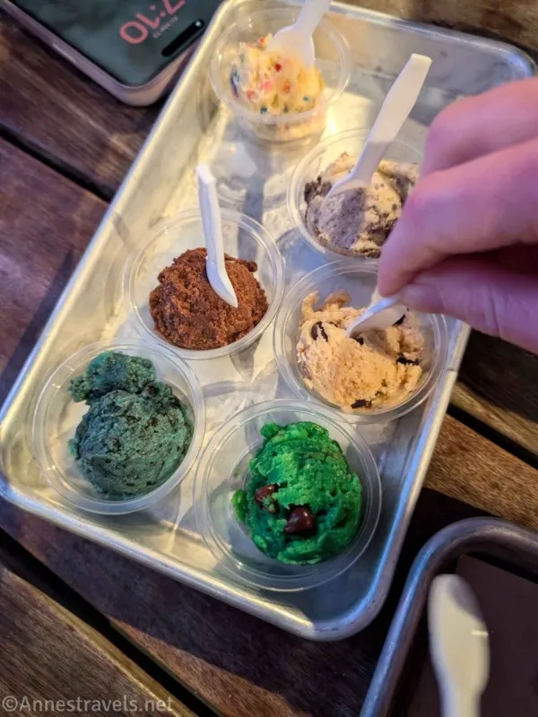 Six cups with colorful cookie dough and three spoons, one of which is held by a hand, on a metal tray on a wooden table 