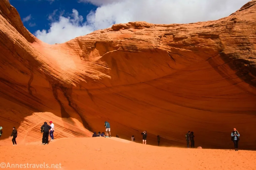 A slickrock alcove with a sand bottom with clouds in the sky above and people in the alcove