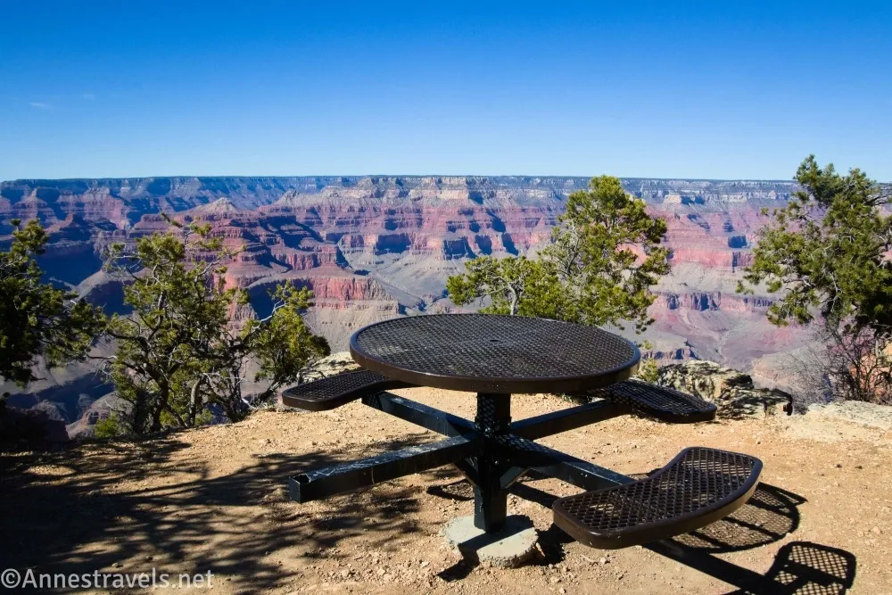 A round picnic table on a gravel area beside trees and the Grand Canyon