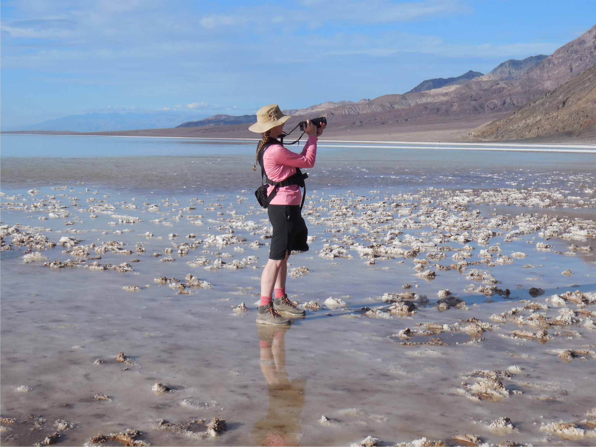 A woman in a pink shirt and black skirt holding a camera stands in a film of water with salt formations poking out of the water with distant mountains