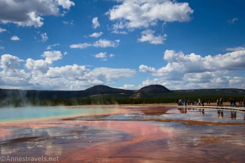 People on a boardwalk beside the colorful Grand Prismatic Spring below puffy white clouds in a blue sky in Yellowstone National Park