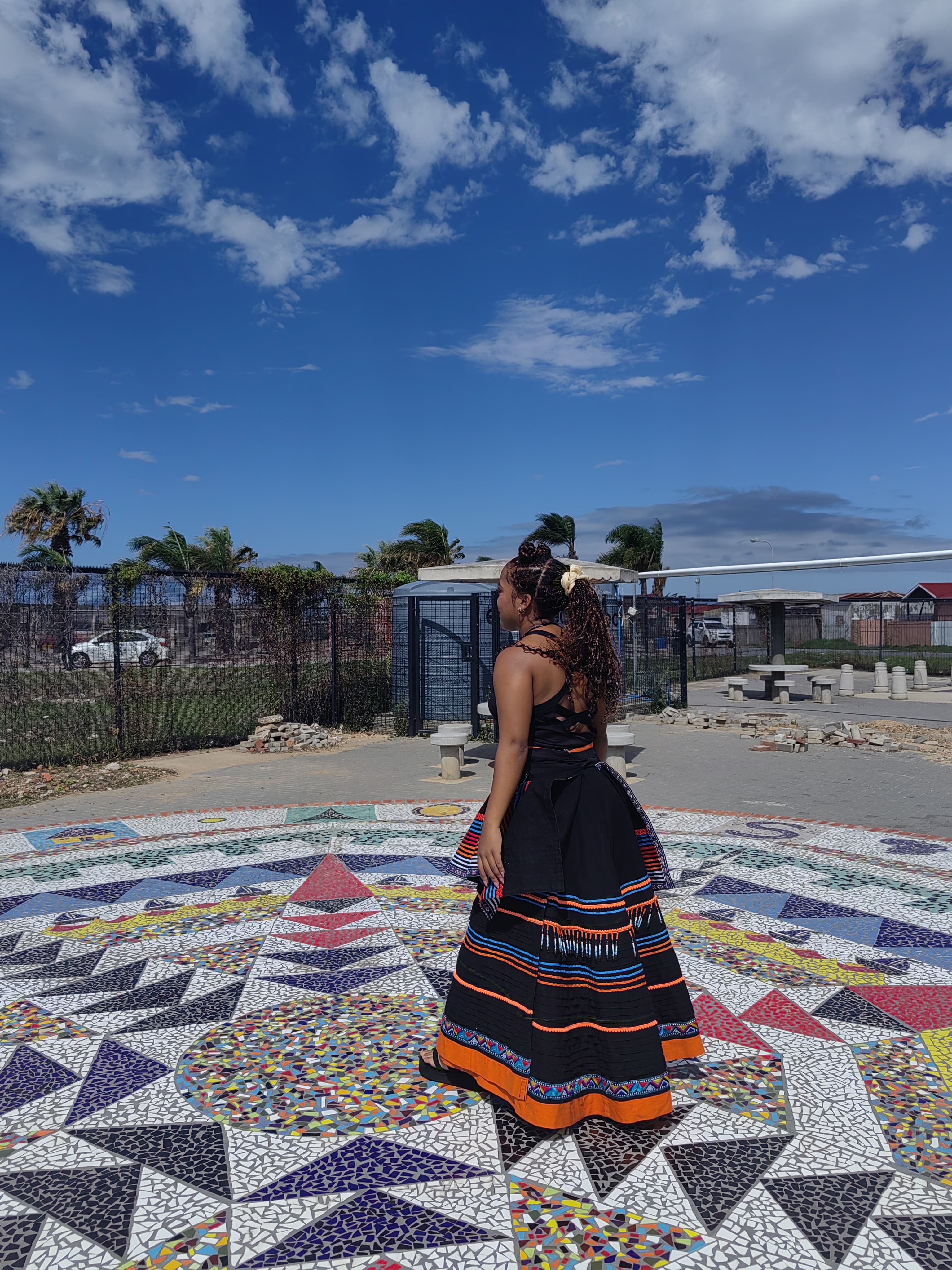 Nanda in a black Xhosa beaded ceremonial skirt on a mosaic mandala, wide sky
