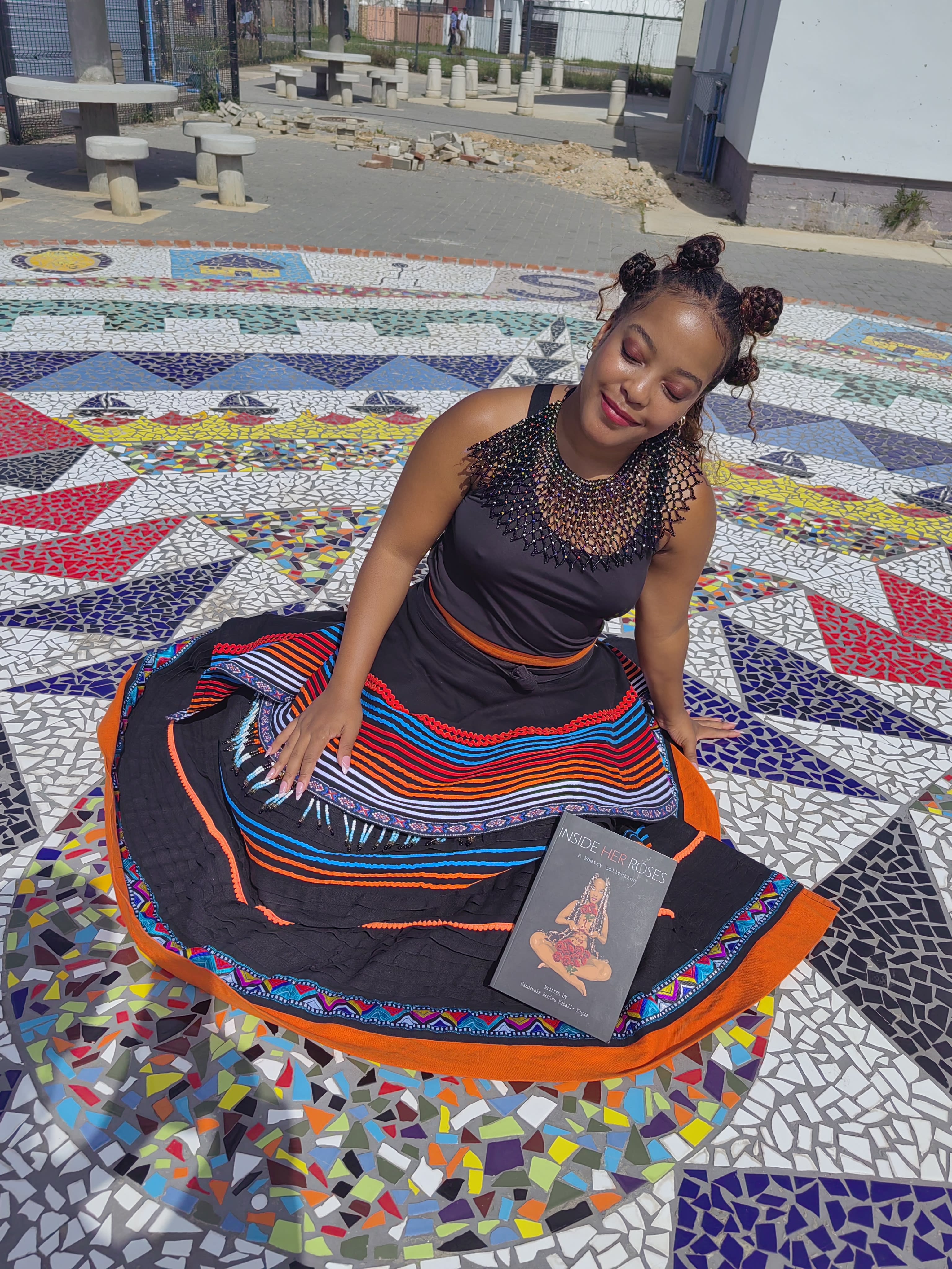 Seated in Xhosa beadwork on a mosaic plaza, holding the book