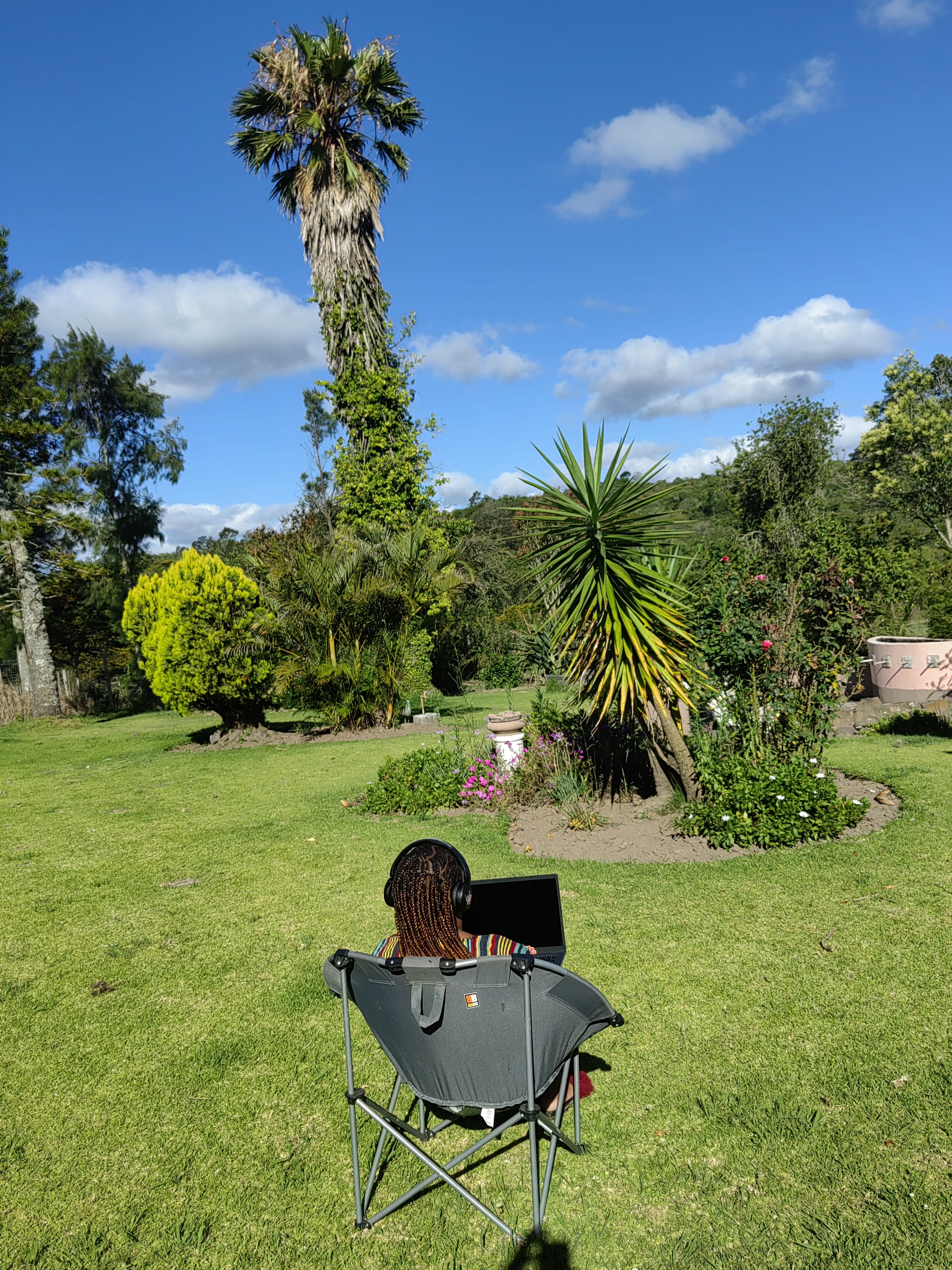 Nanda working from a camp chair on the lawn beneath a palm, laptop on her knees