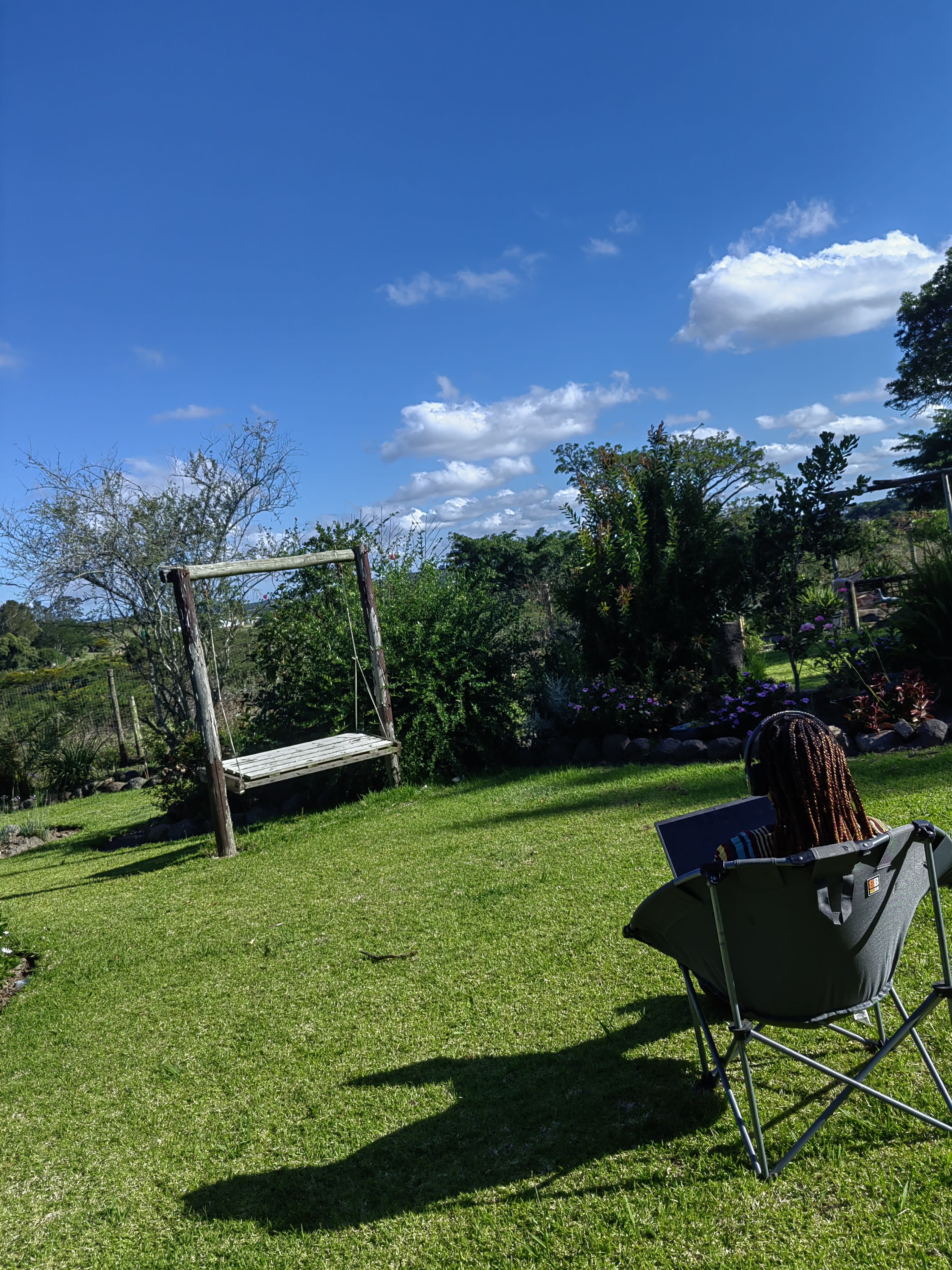 Nanda's laptop open on a table by a garden swing, long afternoon shadows across the lawn