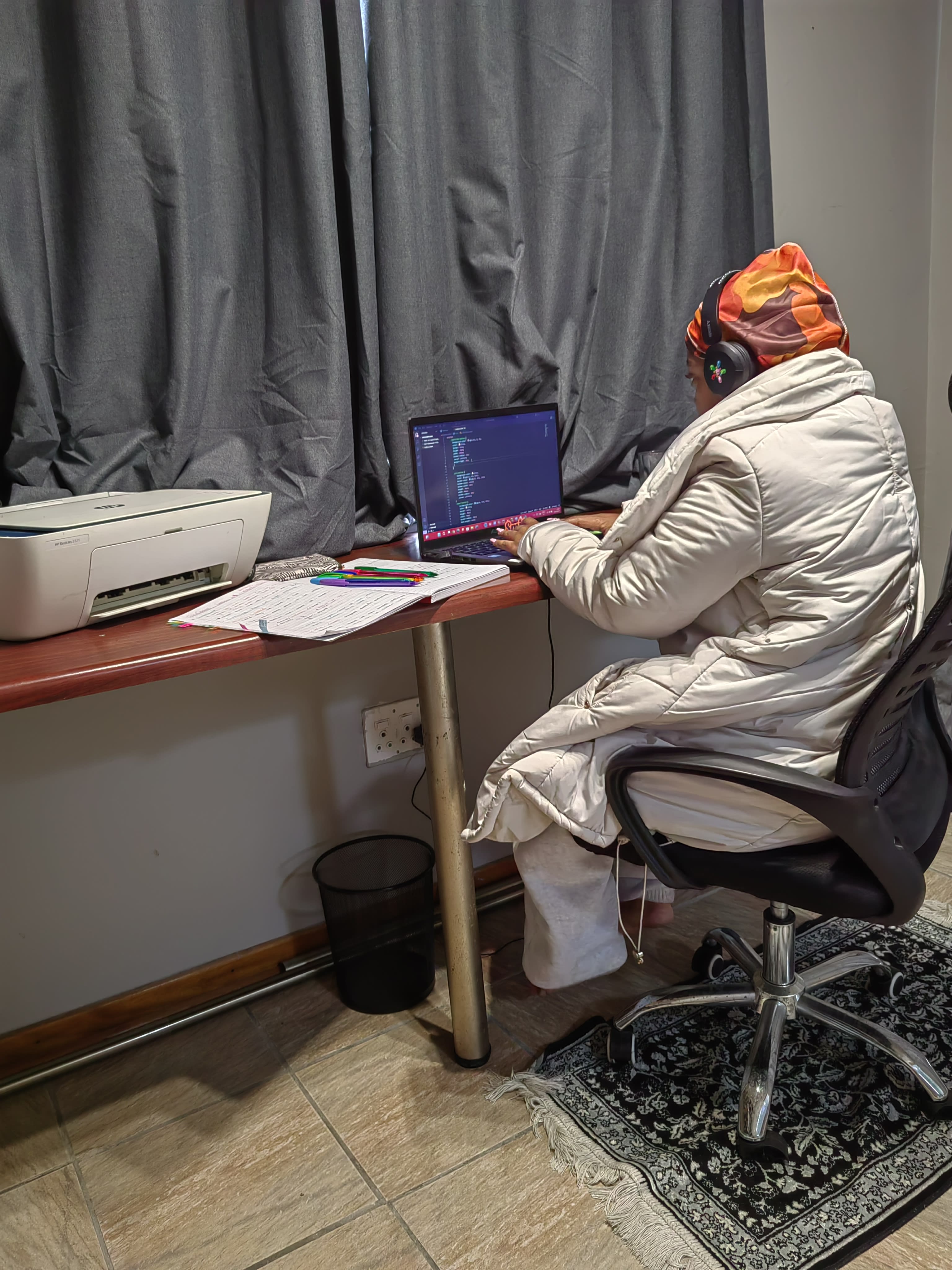 Nandawula Regine at a desk at night, hood up, code on the laptop screen, a printer and notebook beside her