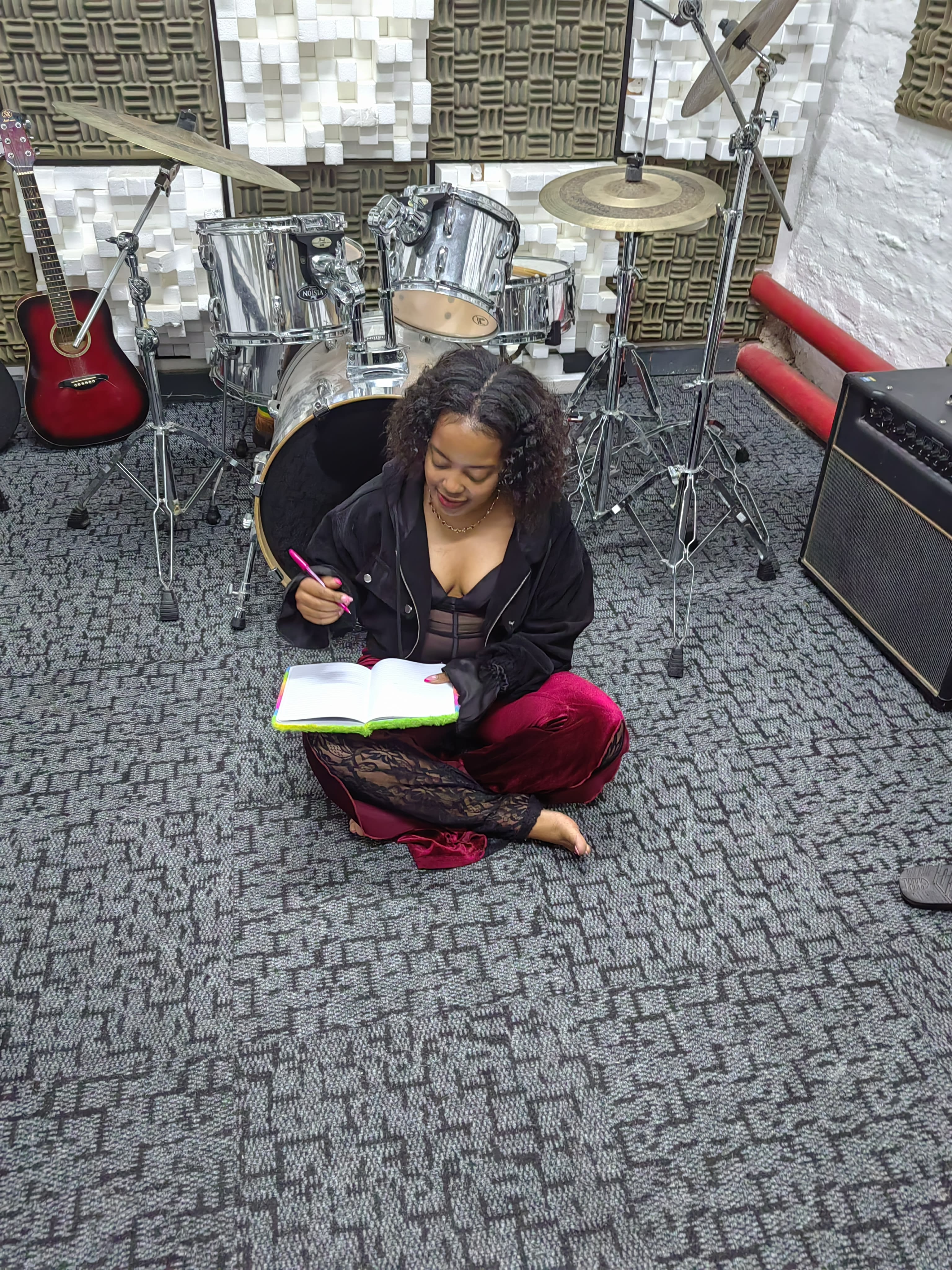 Nanda writing poetry in a notebook, cross-legged among a drum kit and a red guitar