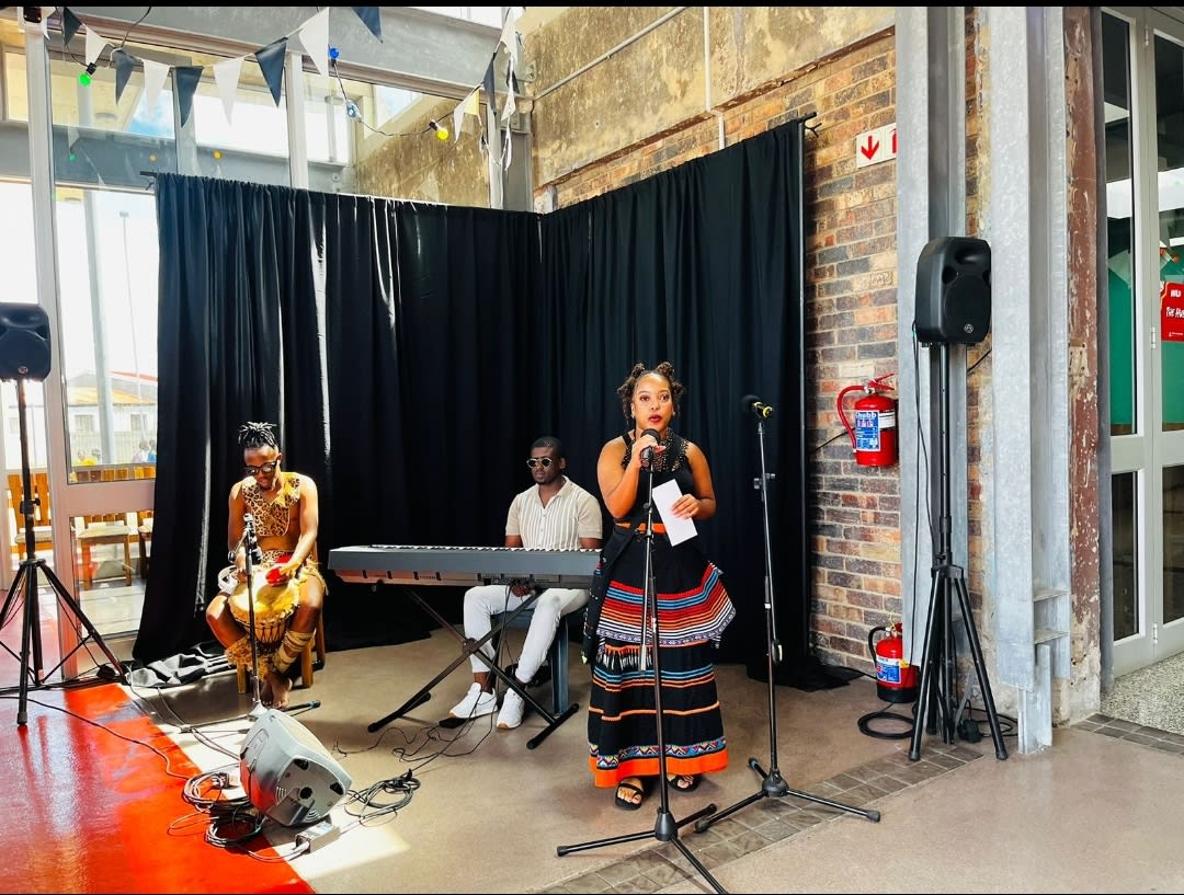 Nanda performing spoken word in Xhosa beadwork with a drummer and keyboardist