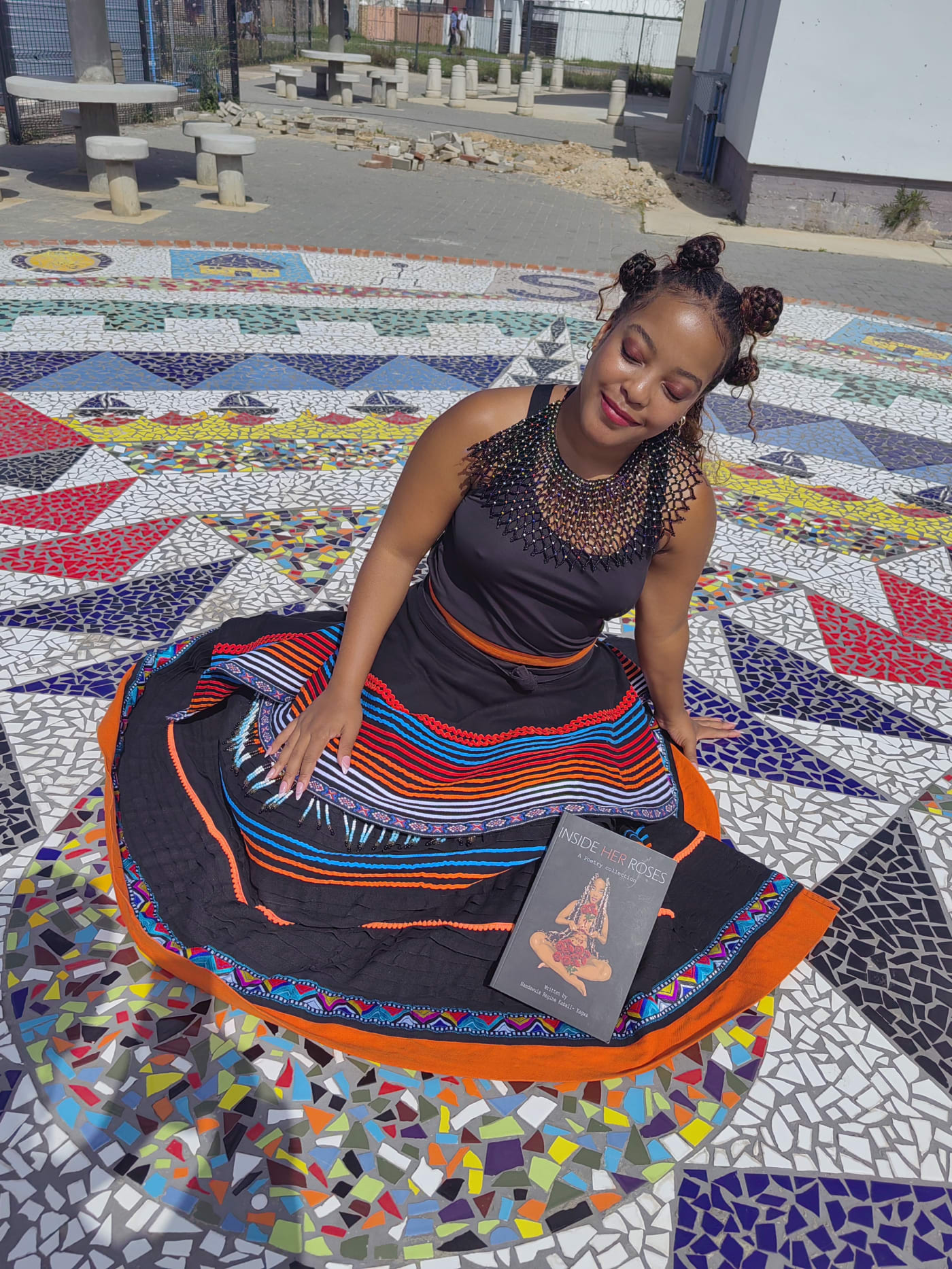 Seated on the mosaic plaza in Xhosa regalia, holding the book