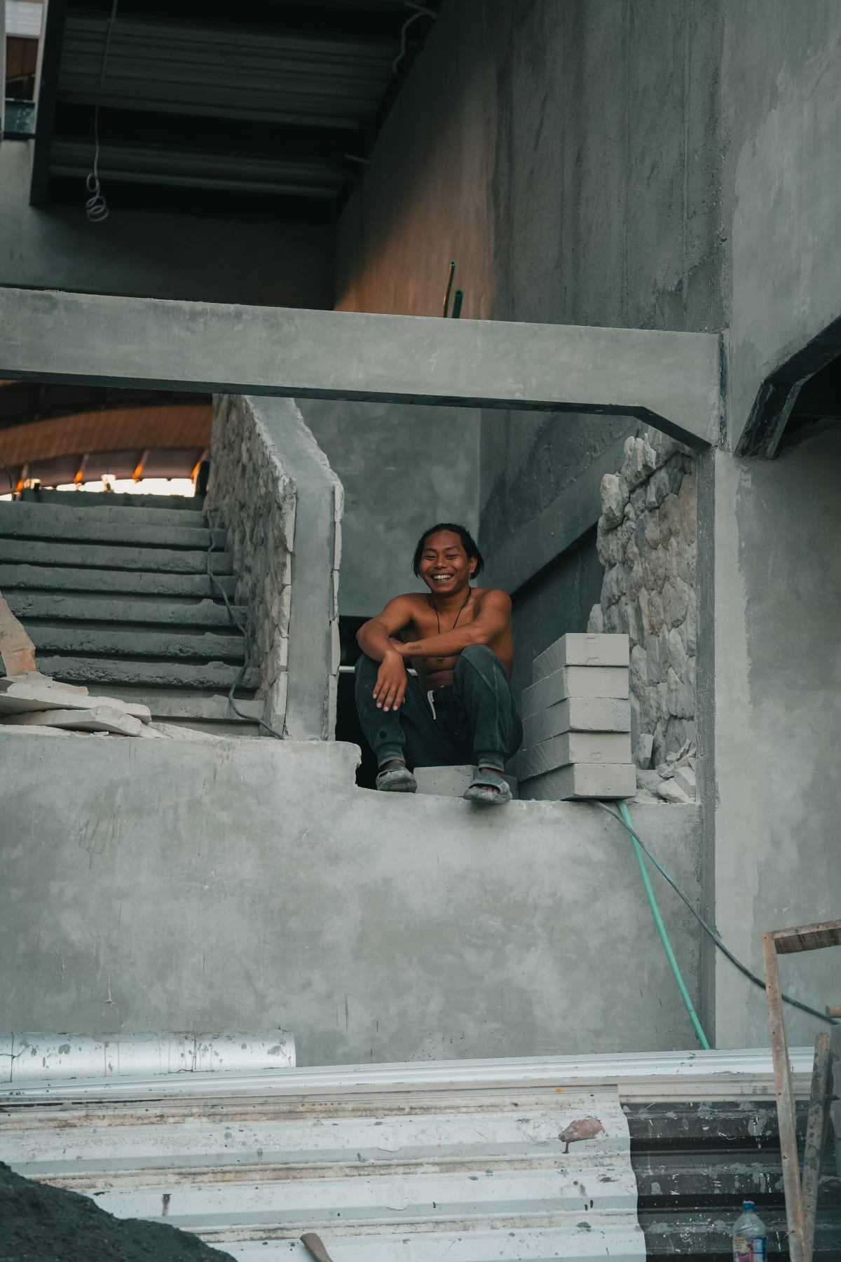 Smiling Balinese construction worker portrait, shirtless, sitting on concrete ledge, unfinished building interior, grey tones, Bali, Indonesia.