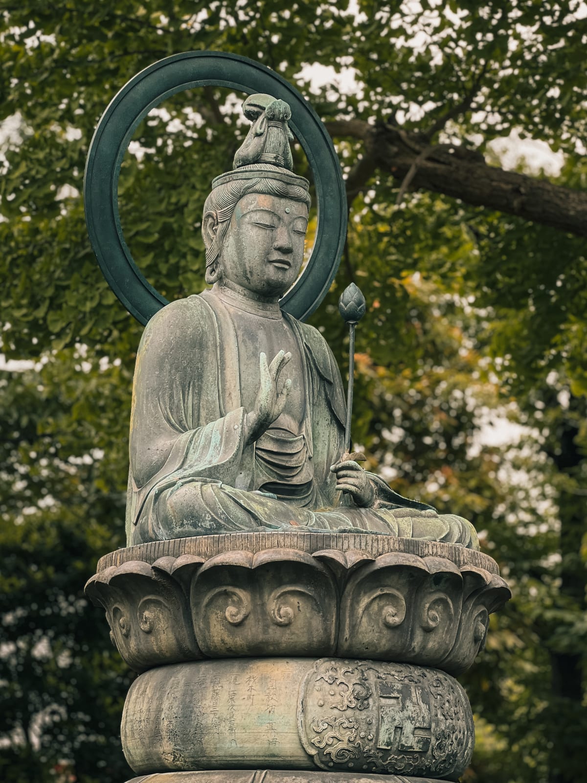 Serene bronze Buddha statue in Taito, Japan. Meditative pose, holding lotus bud, intricate pedestal, lush green temple garden backdrop.
