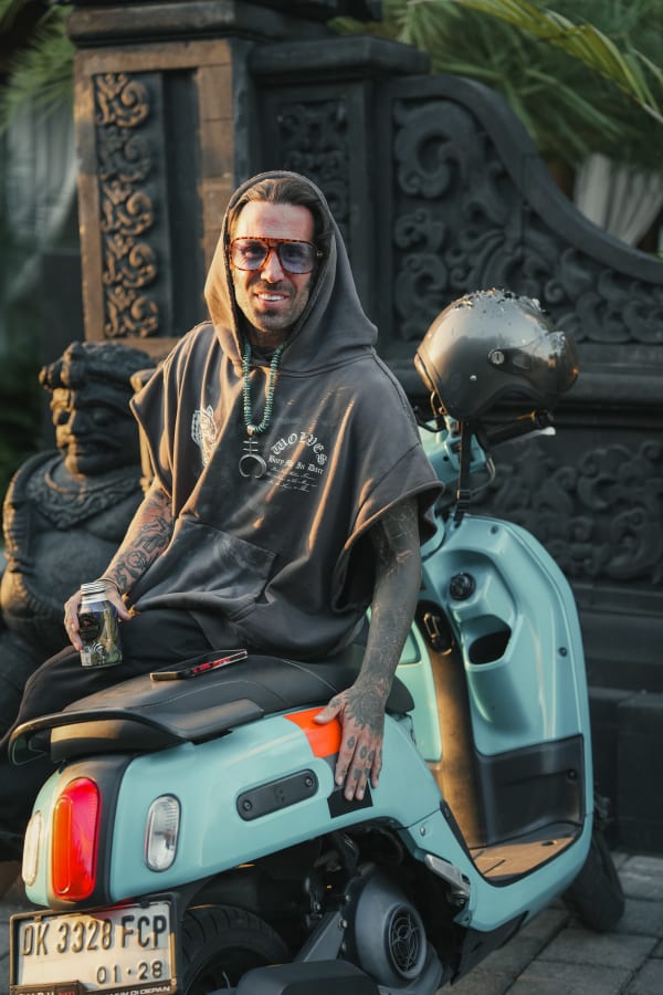 Smiling man on light blue scooter in Bali, wearing hoodie, sunglasses, and beaded necklace, beside ornate Balinese stone carvings at sunset.
