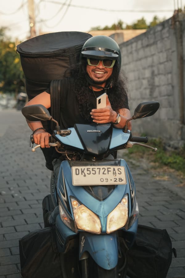 Smiling Indonesian delivery rider on blue Honda Vario scooter in Bali. Man with green helmet, sunglasses, phone, large black bag. License plate DK5572FAS.