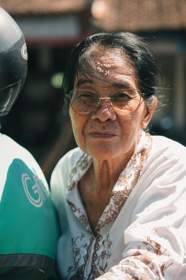 Elderly Balinese woman portrait, glasses, white shirt, rice grains on forehead. Grab uniform visible, Bali, Indonesia.