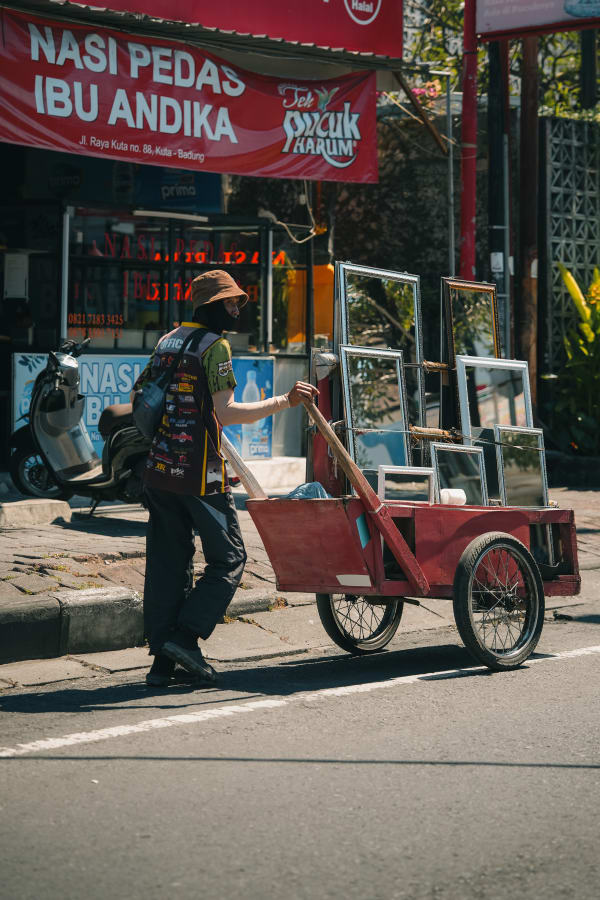 Man in hat pushes red wooden cart with framed mirrors down sunny Kuta street, Bali, Indonesia, past Nasi Pedas Ibu Andika.