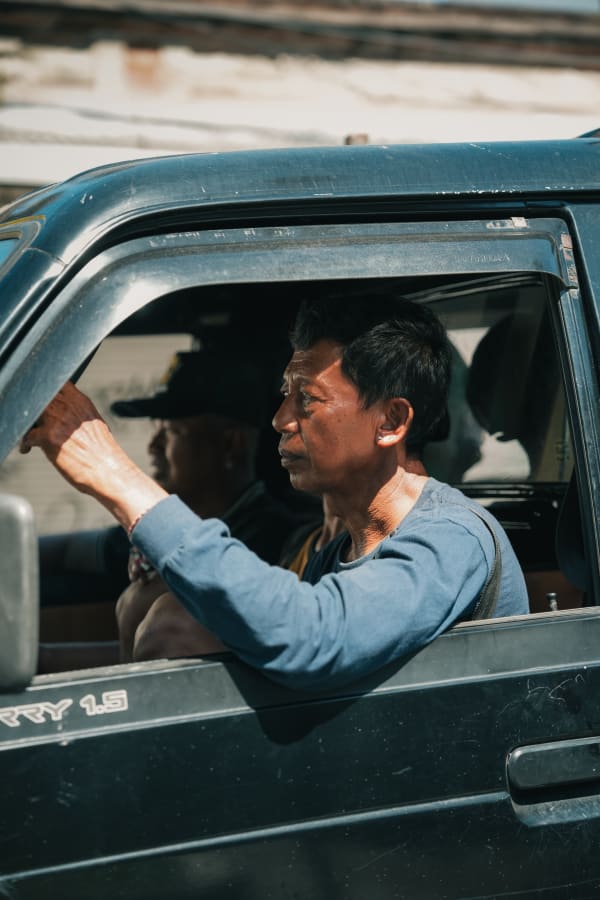 Elderly Balinese man drives Suzuki Carry Futura 1.5 in bright sun, Bali street scene, Indonesia. Side profile.