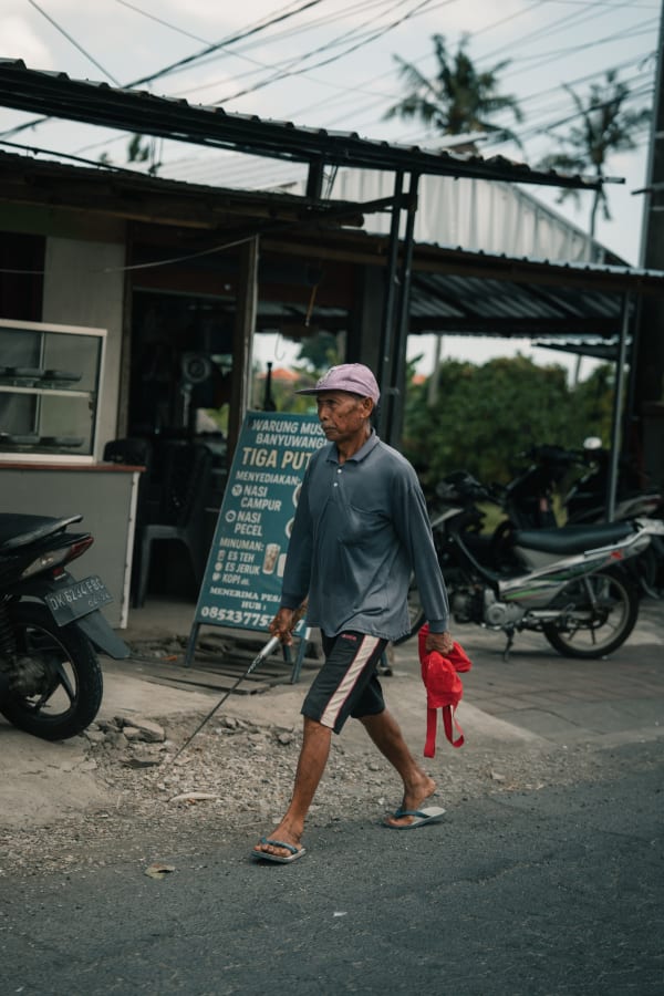 Elderly Balinese man in purple cap walks past a street warung and parked motorbikes, holding a stick and red cloth, Bali, Indonesia.