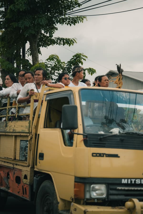 Balinese locals in traditional white attire, some with headbands, riding in an old yellow Mitsubishi truck, Bali, Indonesia.