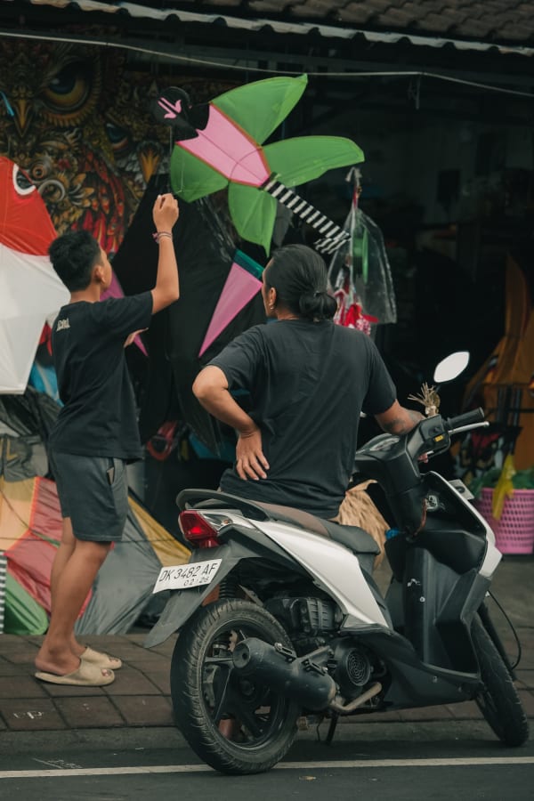 Bali street scene with two men, one on a scooter, observing vibrant green and pink kites hanging in front of a shop with an intricate owl mural.