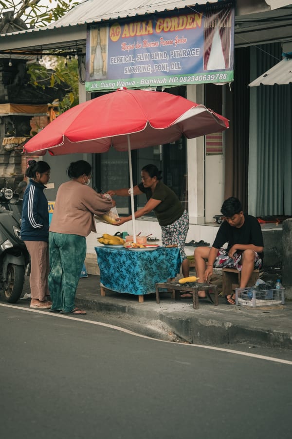 Blahkiuh, Bali street food vendor selling grilled corn under a red umbrella, with customers and a man grilling on the sidewalk.