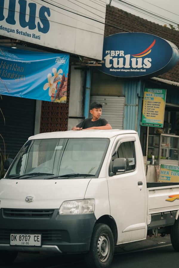 Young boy looks out from white pickup truck on a Bali street, with Indonesian warung sign and Hari Raya Kuningan banner.
