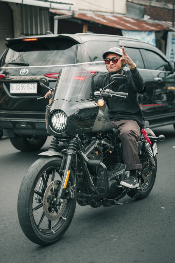 Cool man in cap, red sunglasses on black Harley-Davidson motorcycle, hand gesture, Bali street scene, Indonesia urban portrait.