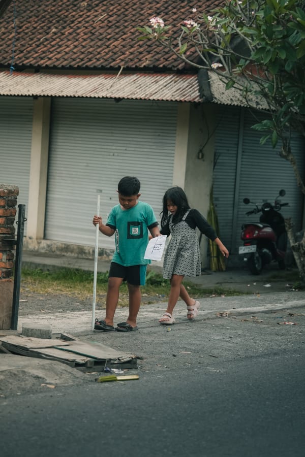 Two Balinese children walk along a quiet street, boy with stick, girl with paper, past closed shops and a frangipani tree in Bali.