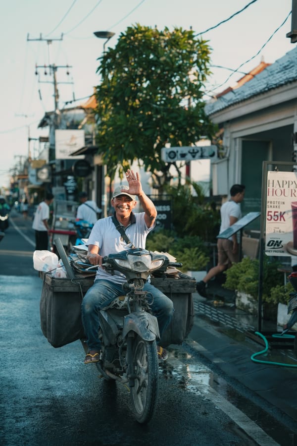 Smiling Balinese man on rustic motorcycle, carrying goods, waving on bustling street, golden hour, Bali, Indonesia.