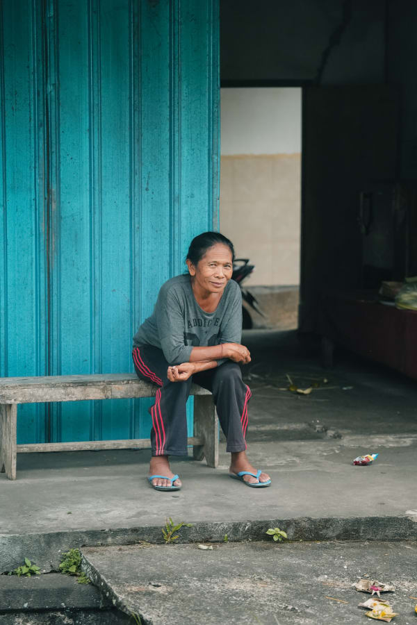 Balinese woman on wooden bench in front of turquoise door in Bali, Indonesia, with gentle smile, local life street scene.