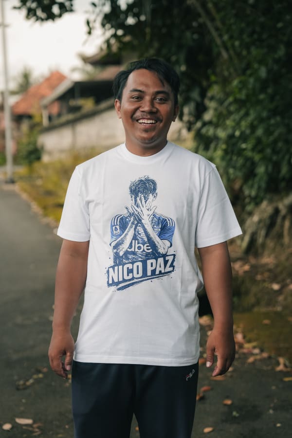 Smiling Balinese man in white 'Nico Paz' t-shirt on a village street in Bali, Indonesia, with blurred foliage.