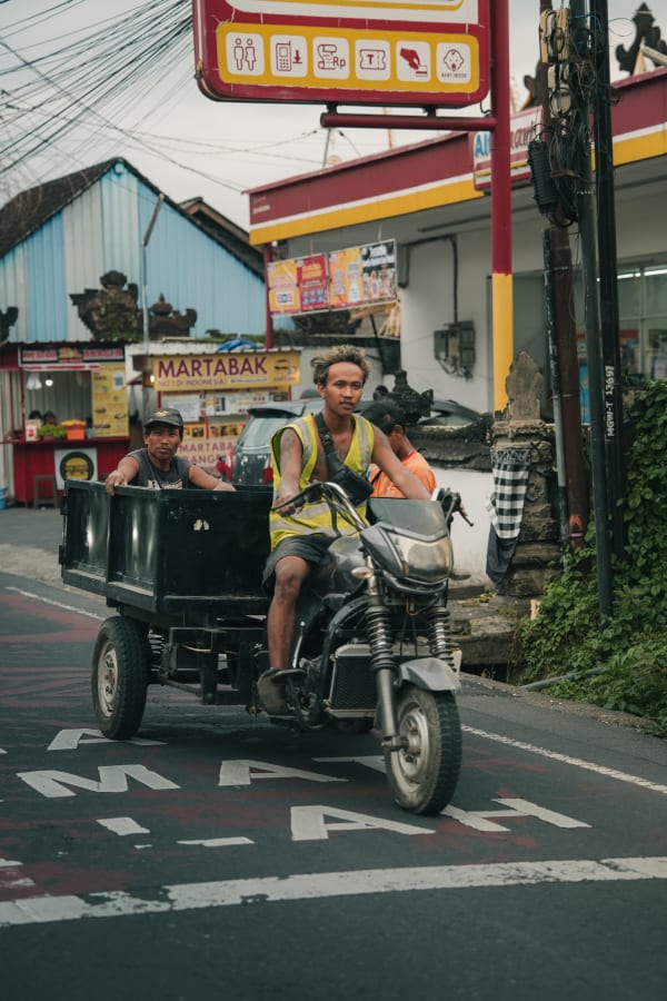 Two men on a utility scooter with a trailer pass a Martabak stall on a vibrant Balinese street, Indonesia, under overcast skies.