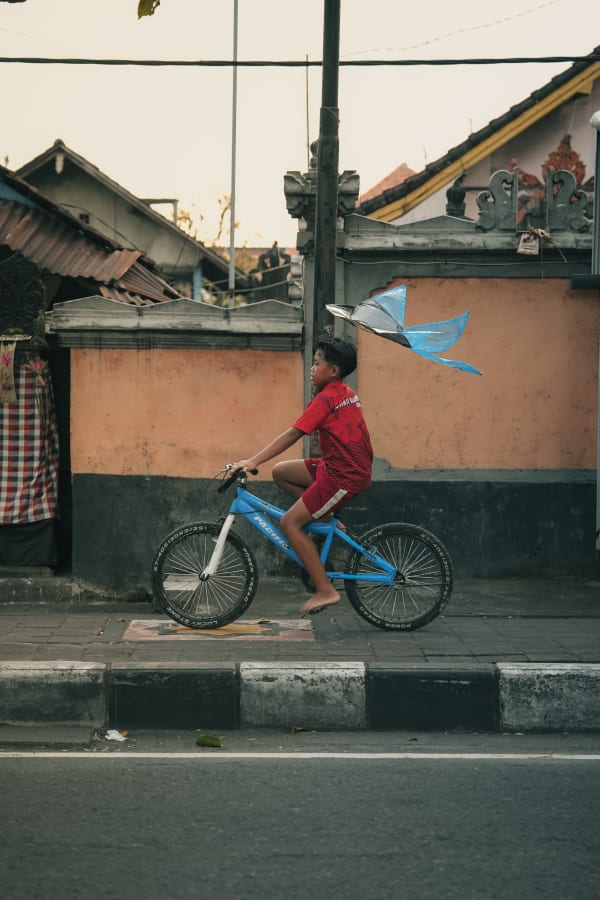 Barefoot Balinese boy on blue bicycle with kite, vibrant red shirt, riding past traditional Balinese house wall, soft light, Bali, Indonesia.