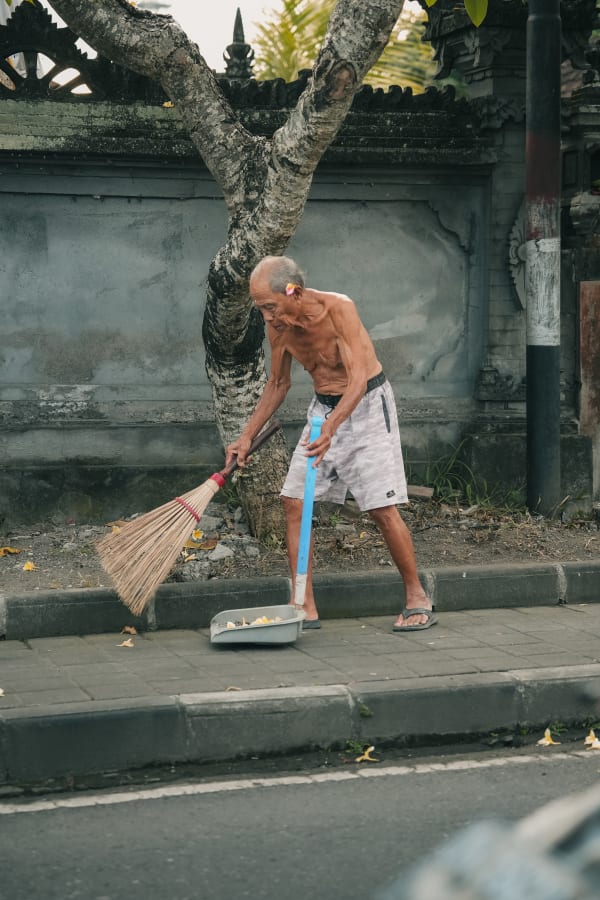 Elderly Balinese man sweeping a street in Bali, Indonesia, with a frangipani flower behind his ear, by a traditional carved stone wall.