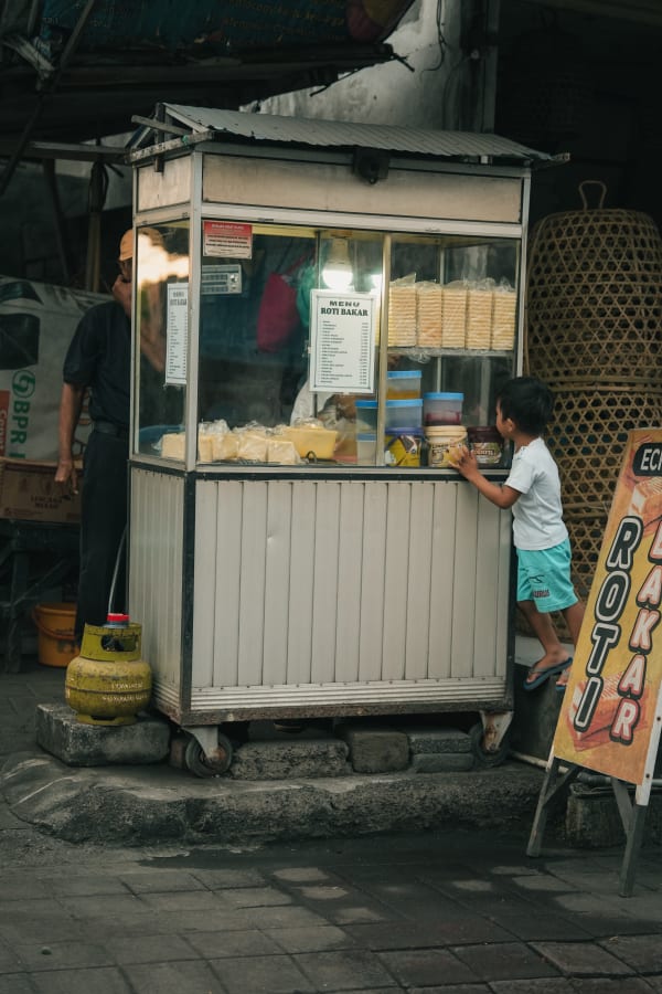 Child, vendor, Roti Bakar street food cart in Bali. Indonesian urban scene, a yellow gas tank sits beside.