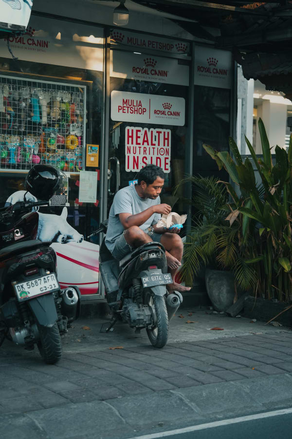 Man barefoot eating on a scooter in front of Mulia Petshop, Bali. Tropical street scene with Royal Canin signs, green plants, soft light.